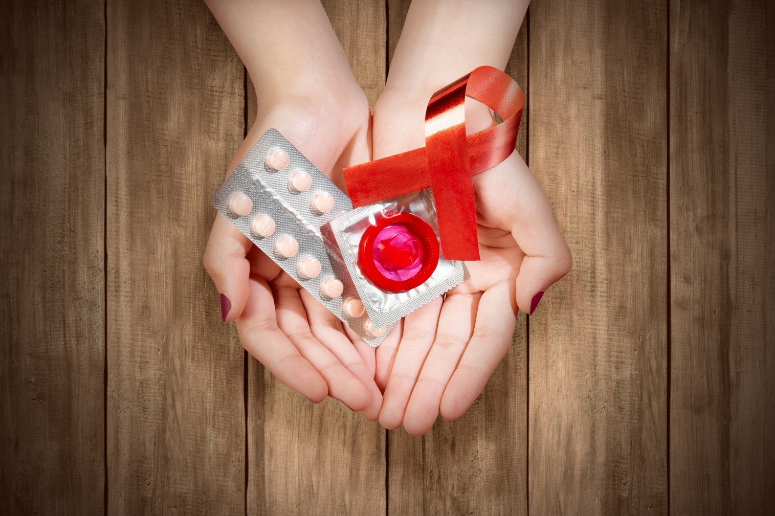 Photo of hands holding a red ribbon, condom and pill packet on a wooden background