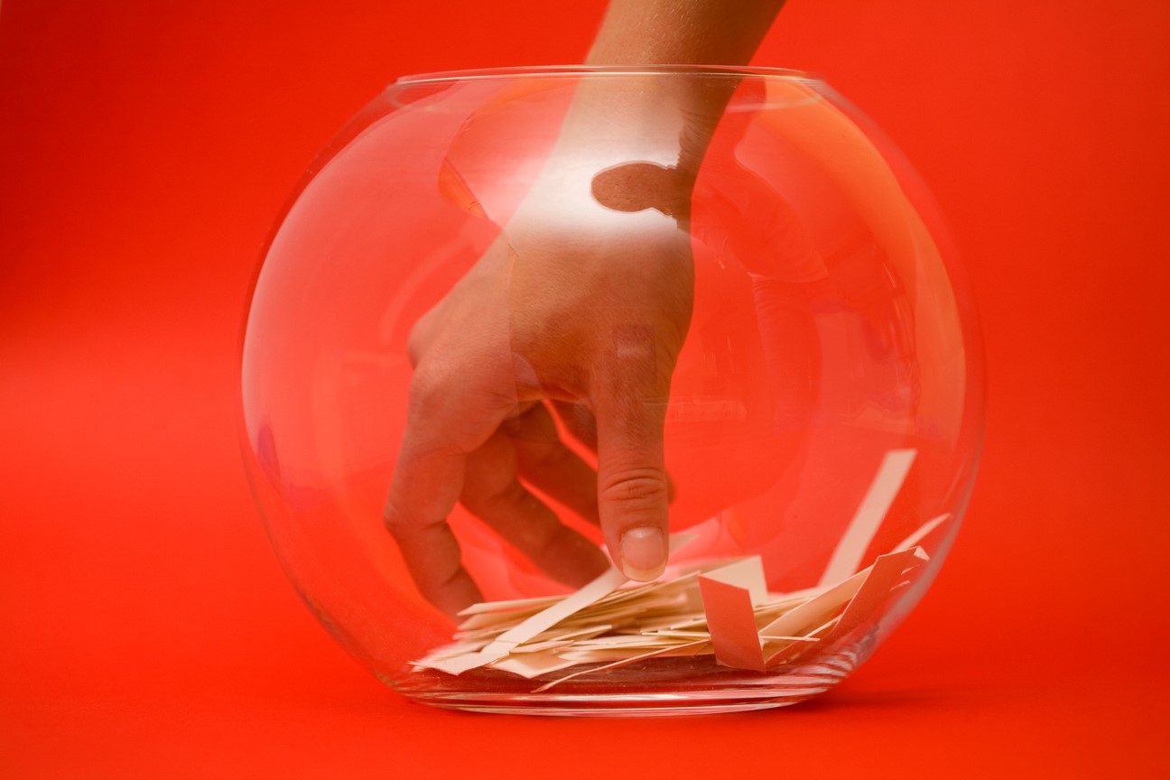 Photo of a hand reaching into a glass bowl with small paper slips, in front of a red background