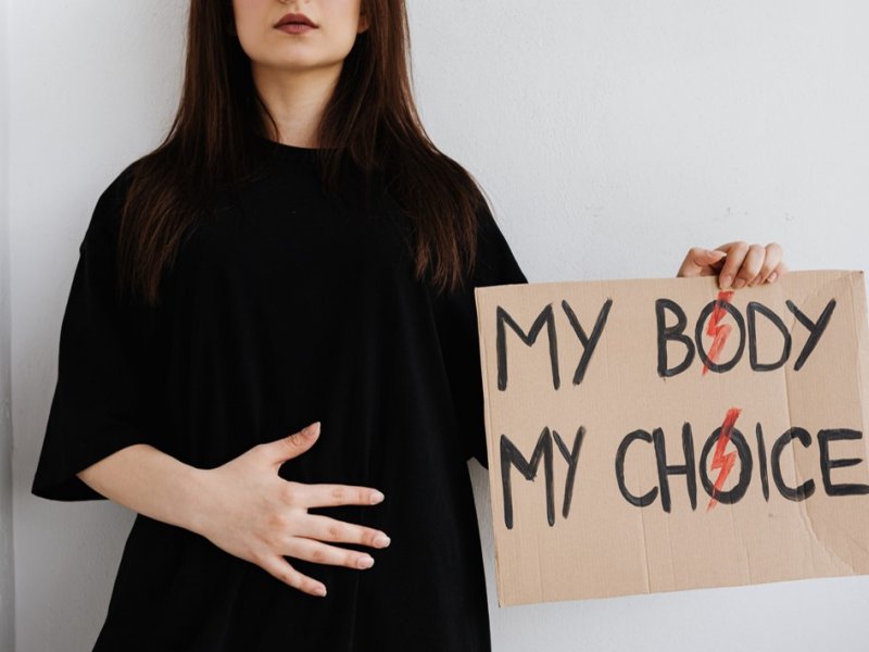 Photo of a person in a black smock holding a sign that says "My Body My Choice"